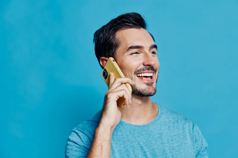 Smiling man using smartphone while talking on the phone, dressed in casual blue t-shirt, cheerful expression, isolated on vibrant blue background. People lifestyle concept