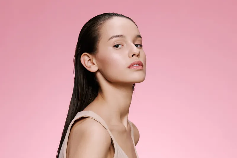 Young woman with long wet hair posing confidently against a pink background, showcasing beauty and elegance in a minimalistic style with a soft color palette