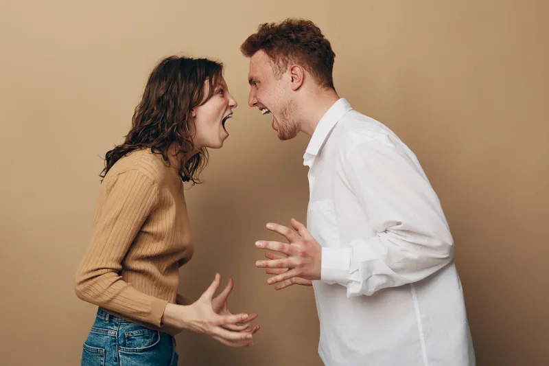 Angry young man and woman shouting at each other with intense expressions. Conflict and argument concept in casual clothing against plain beige background. Emotional tension and disagreement.