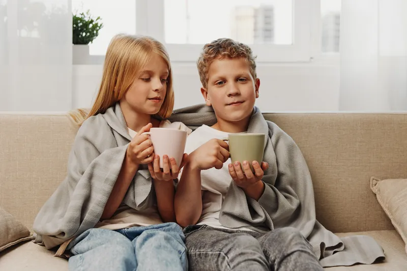 Two children wrapped in blankets sit on a couch holding mugs, enjoying a cozy moment indoors with natural light from windows.