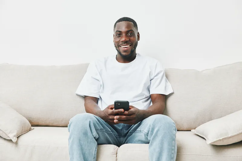 Smiling Black man sitting on a beige sofa, using a smartphone, joyfully engaging with digital content in a bright living space