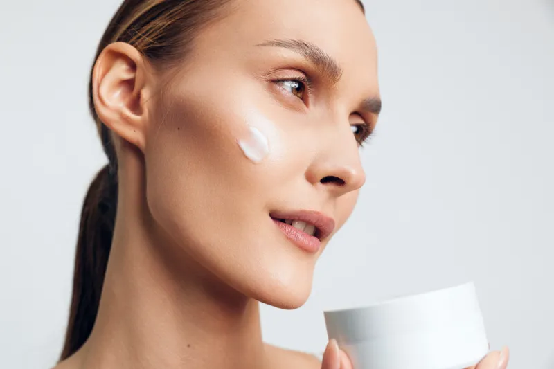 Young woman with clean skin applies white moisturizing cream on her cheek holding white jar close up beauty skincare concept isolated on light background with natural makeup.