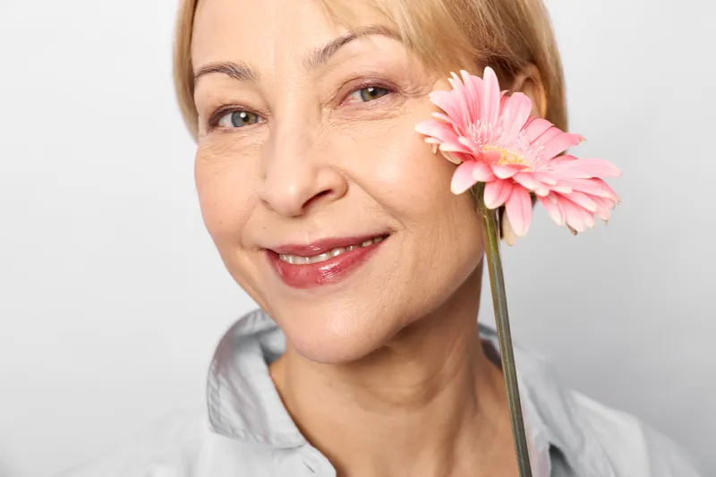 Smiling senior blonde woman in closeup portrait, pink flower beside her ear, soft light and white background, beauty and wellness concept, gentle calm expression and elegance