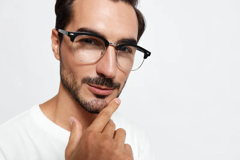 Confident young man with glasses thoughtfully touching his chin while looking at the camera, dressed in a white t-shirt, isolated on a plain light background.