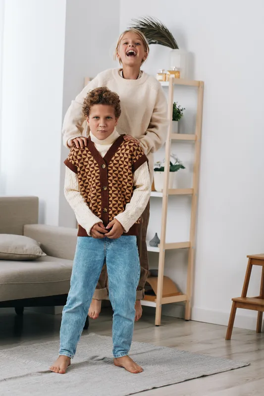 Two smiling children stand indoors as a girl jumps behind a boy in a bright living room. Cozy sweaters, warm light, and carefree energy capture a joyful, candid moment.
