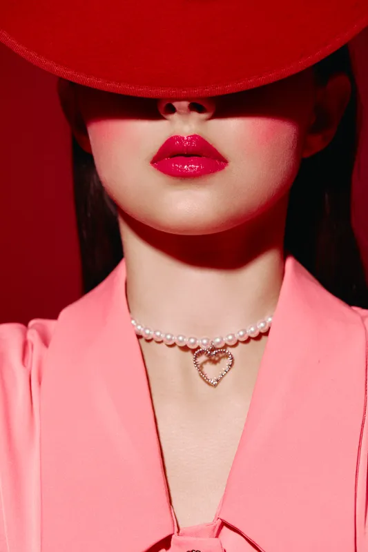 Close-up of a woman with bright red lipstick wearing a pearl choker with heart pendant and a pink jacket. Shadowed eyes create a mysterious fashion beauty concept.