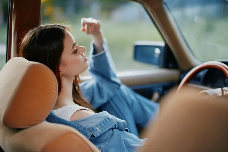 Young woman in denim jacket, relaxing in vintage car, exuding calmness and nostalgia in a lush green background, emphasizing freedom and adventure.