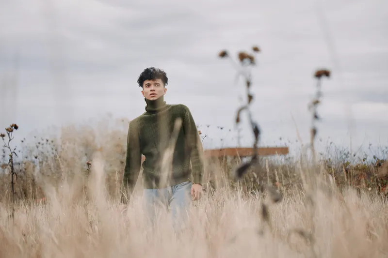 Young man in a green turtleneck stands in tall dried grass under an overcast sky, looking thoughtful and present; an authentic portrait capturing credible, natural emotion and simple rural texture.