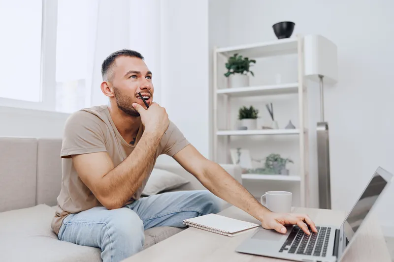 Thoughtful man working on a laptop in a modern living room, reflecting on ideas while enjoying a cozy atmosphere. Perfect for illustrating productivity and creativity.