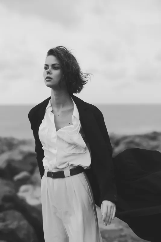 Elegant fashion on a windy coast featuring a woman in a white shirt, blazer and trousers, captured in monochrome with a flowing coat by the seaside and a confident stance.