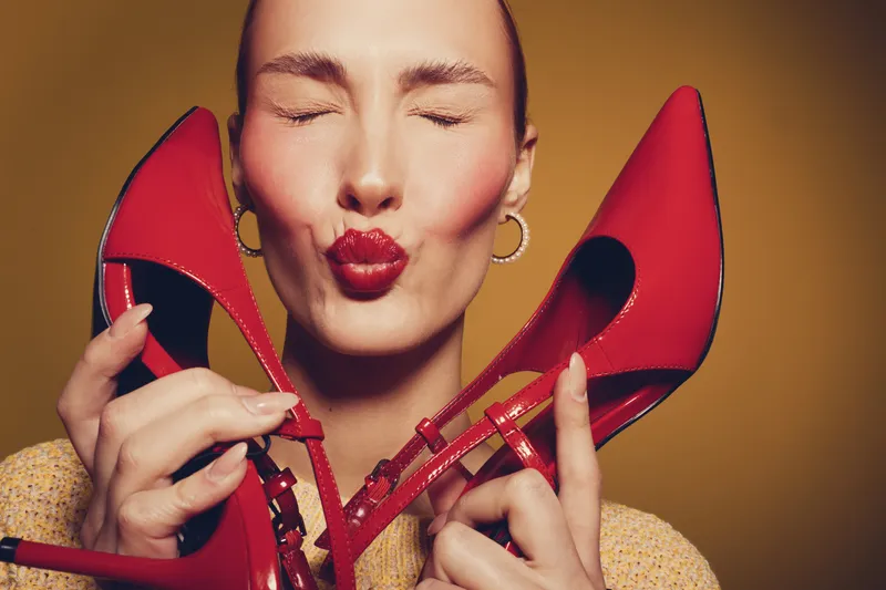 Close-up of a happy woman holding red high heels near her face with closed eyes and puckered lips. Concept of fashion, style, footwear, and joyful emotions on warm background.