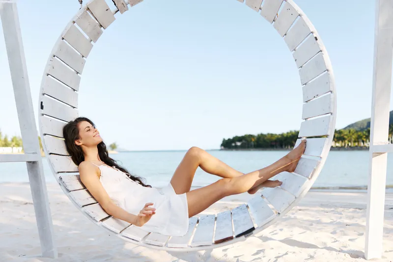 A carefree woman relaxing on a circular white swing by a sunlit beach, wearing a breezy white dress, with calm sea and clear blue sky in the background.