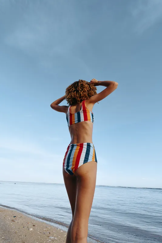 Back view of a woman in a colorful striped swimsuit standing on a sandy beach, watching the calm sea under a blue sky, capturing a sunny summer moment