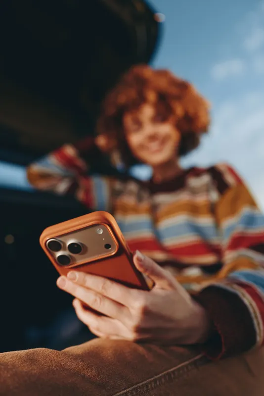 Curly woman in striped sweater uses smartphone on sofa at home lifestyle, hands holding phone for texting and communication in relaxed evening warm light
