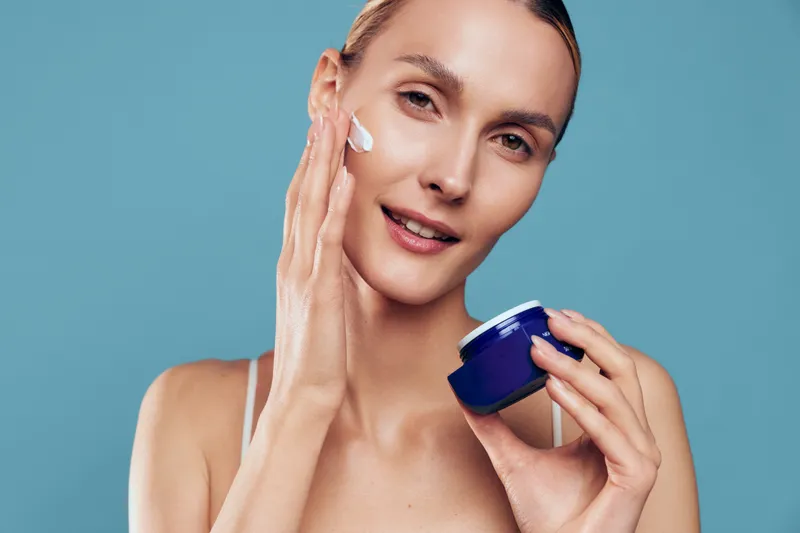 A young woman with clear skin applies moisturizing cream to her face while holding a blue jar against a solid light blue background. Concept of skincare and beauty routine.