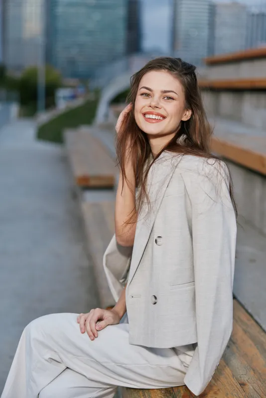 Young smiling woman in light business suit sitting on city bench, confident woman, outdoor urban setting, modern fashion style, natural emotions