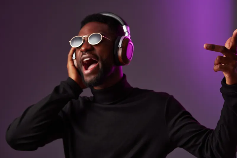 Enthusiastic young man enjoying music with headphones in stylish attire against a purple background evokes passion and joy in contemporary fashion