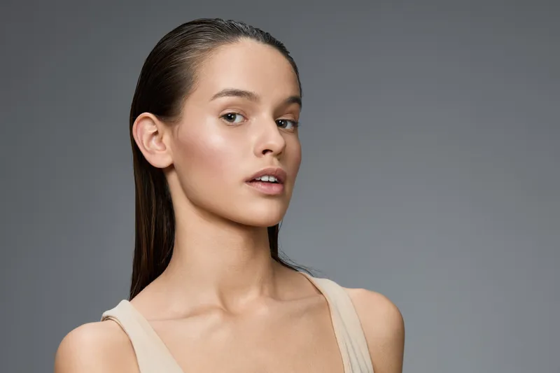 Natural beauty portrait of a young woman with minimal makeup, showcasing healthy skin and long hair against a smooth grey background, emphasizing elegance and simplicity