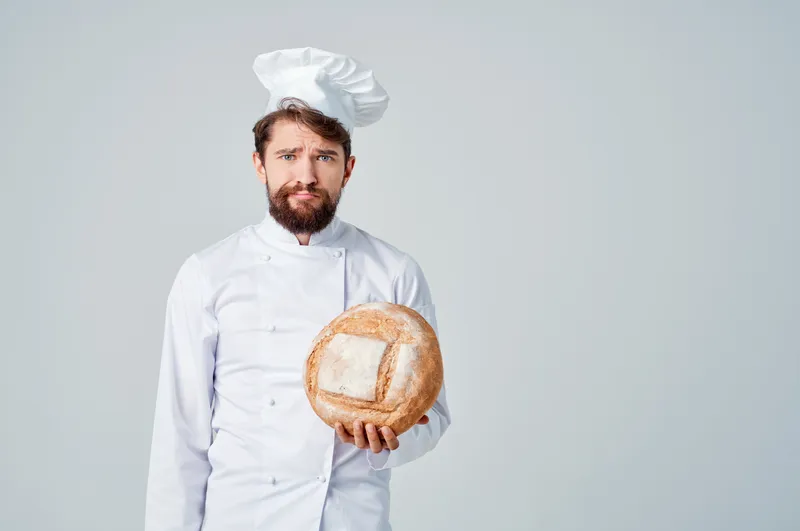 Бородатый мужчина-шеф-повар на кухне, рядом с хлебобулочными изделиями, на светлом фоне