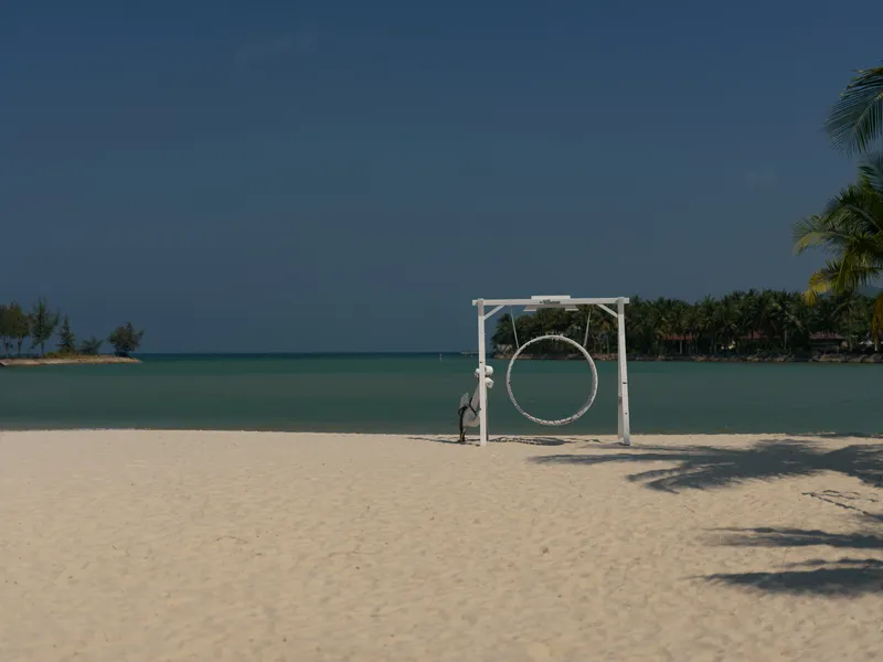 empty sandy beach with a large white circular swing frame near the shore, calm turquoise sea and clear blue sky in the background, tropical vacation setting