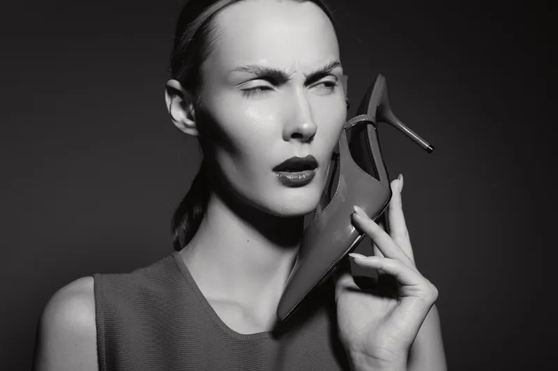 Caucasian woman with pale skin holds high heel shoe to ear in black and white studio portrait. Concept for fashion, style, and expression.