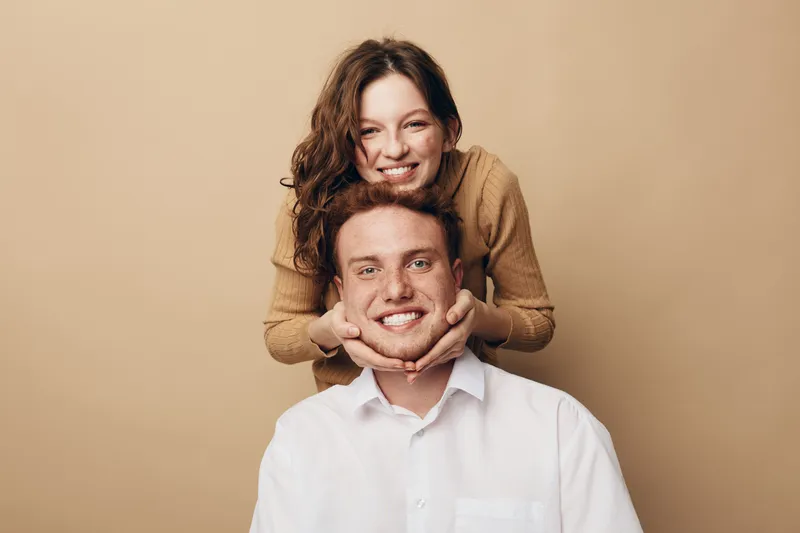Smiling young woman in brown sweater holds happy man in white shirt by chin. Friendly couple poses on beige background. Concept of love, relationship, happiness, and togetherness.