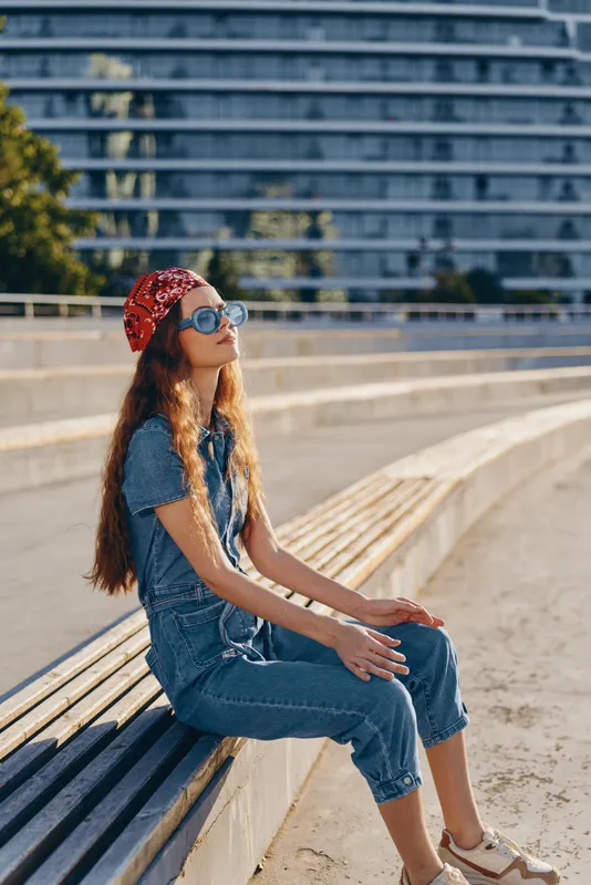 lifestyle woman in modern boho-western denim street style wearing sunglasses and red bandana sitting outdoors with film color effect under bright sunlight