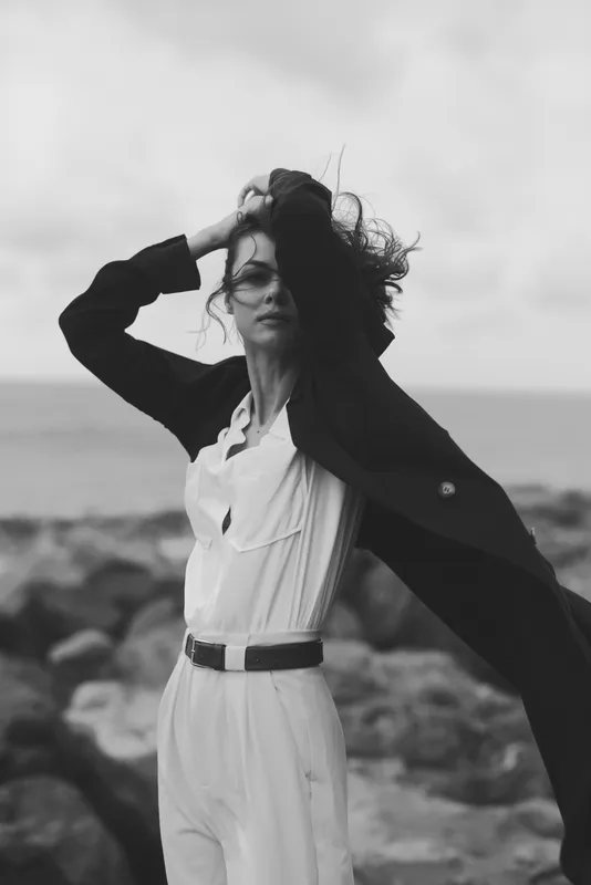 Fashion, model on a windy seaside, flowing white dress with a black coat, dramatic motion by the shore, monochrome mood for editorial storytelling