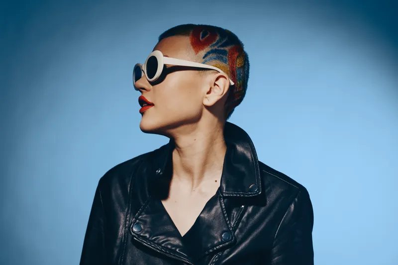 Woman with colorful shaved hair wearing round white sunglasses and black leather jacket, looking sideways against soft blue background, modern style.
