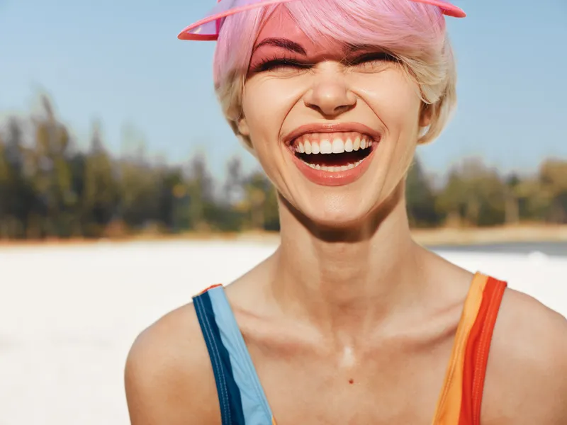 Closeup portrait of a joyful woman with pink hair and pink sunglasses atop her head, laughing under bright sun on a sandy outdoor setting, exuding carefree energy.