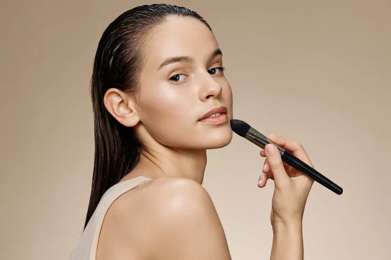 A beautiful young woman applying makeup with a brush against a soft beige background, showcasing a natural beauty concept and glowing skin