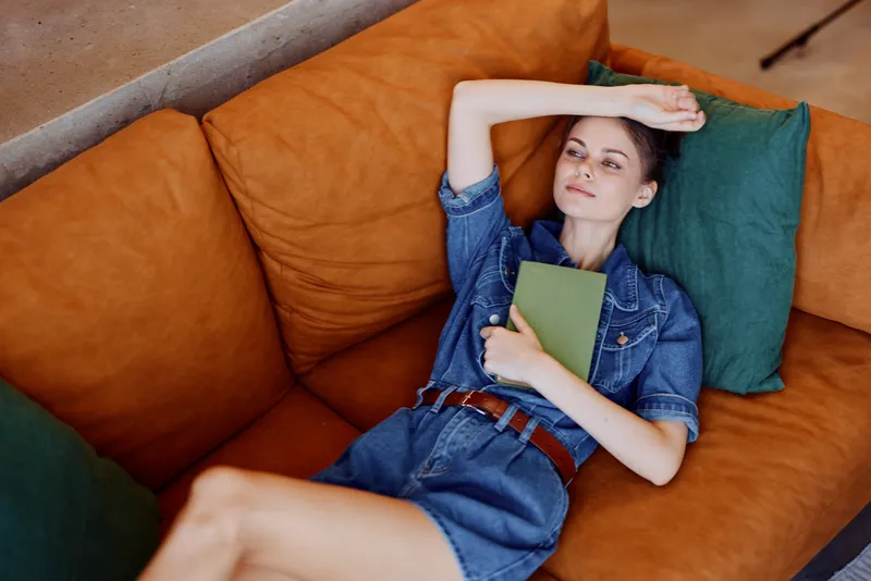 Женщина отдыхает дома, лёжа на диване с книгой, положив голову на подлокотник, — уютный и умиротворённый момент чтения
