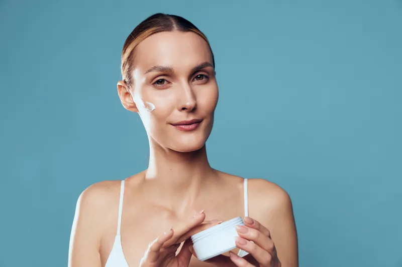 Young woman with clear skin applying white facial cream on cheek, holding jar in hand, wearing white top, isolated on blue background, concept of skincare and self-care routine.