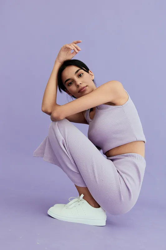 young woman in casual lavender outfit crouching against purple background, wearing white sneakers with natural makeup and relaxed expression, minimalistic style