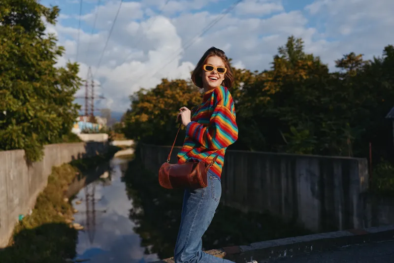 Woman in a colorful sweater and sunglasses, wearing denim jeans and carrying a brown leather bag, smiling by an urban canal framed by autumn trees in golden light, mid twenties joyful traveler.