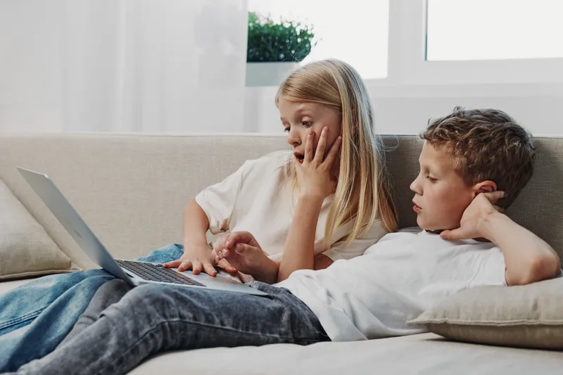 Two children lying on a couch using a laptop together, focused on the screen in a bright living room. Concept of technology, childhood, learning, and sibling interaction.