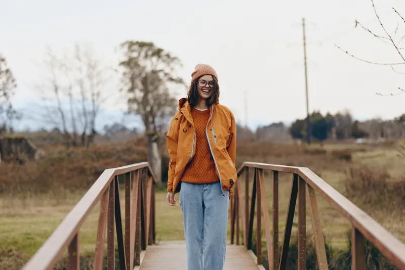Woman lifestyle outdoors walking on wooden bridge in autumn, wearing orange jacket and blue jeans, smiling at camera, soft natural light, rural landscape, calm peaceful mood.