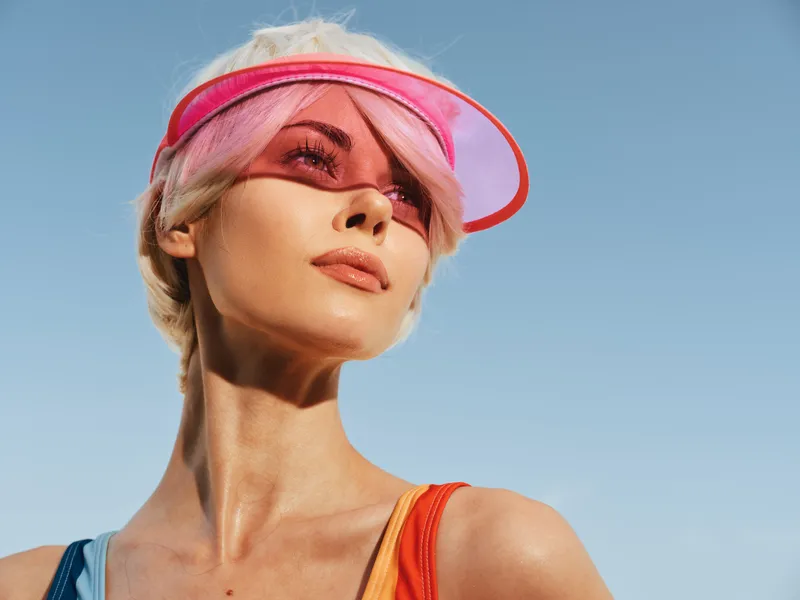 Portrait of a stylish woman wearing a pink visor under a clear blue sky, sunlit skin, and chic summer fashion in a candid outdoor pose.