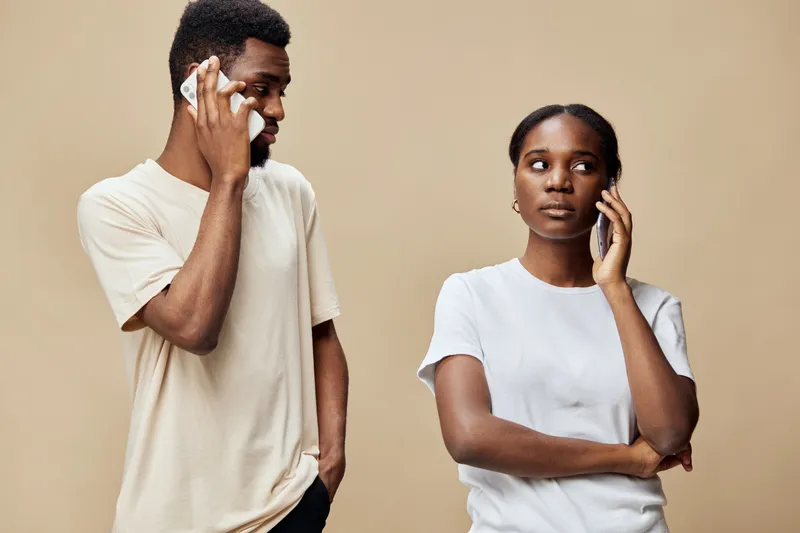 A thoughtful black couple engaging in a conversation, with one holding a smartphone, conveying emotions of contemplation and connection, against a neutral beige background
