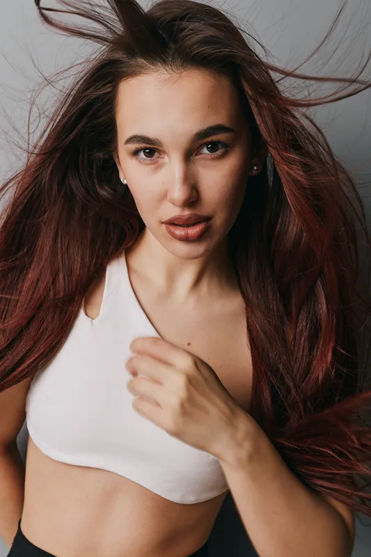 Young woman with long hair wearing white crop top looking at camera with confident expression indoors. Portrait of female model with natural makeup and casual style in studio setting.