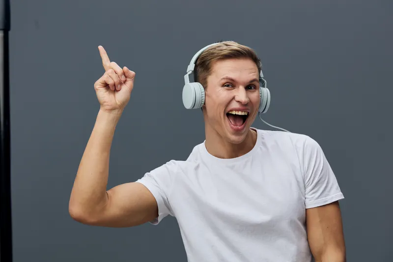 Joyful young man wearing white t-shirt and headphones, expressive happy face, pointing upwards with finger, minimalistic grey background, studio shot, lifestyle concept.