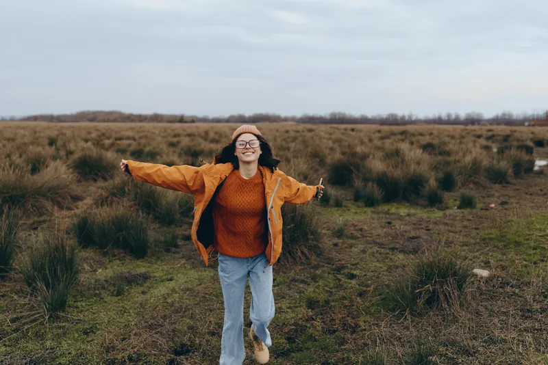 Smiling woman outdoors in autumn field, walking forward with arms open, hat orange jacket and glasses, nature grass landscape, casual lifestyle, wide view, calm mood
