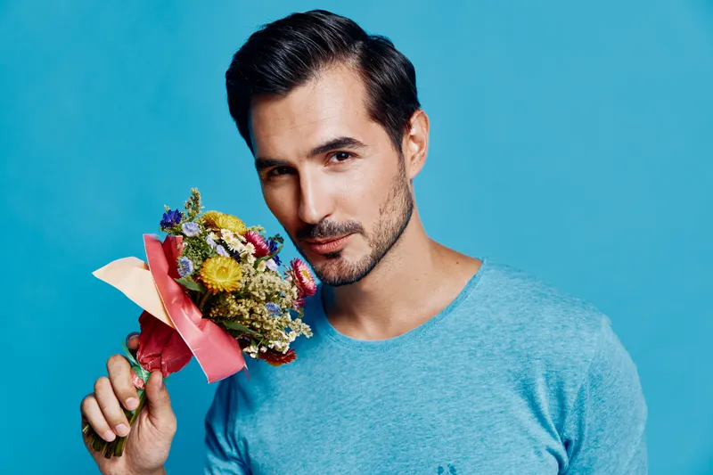 Handsome man holding a colorful bouquet of flowers, smiling gently at the camera against a bright blue background. Positive emotion and lifestyle concept.