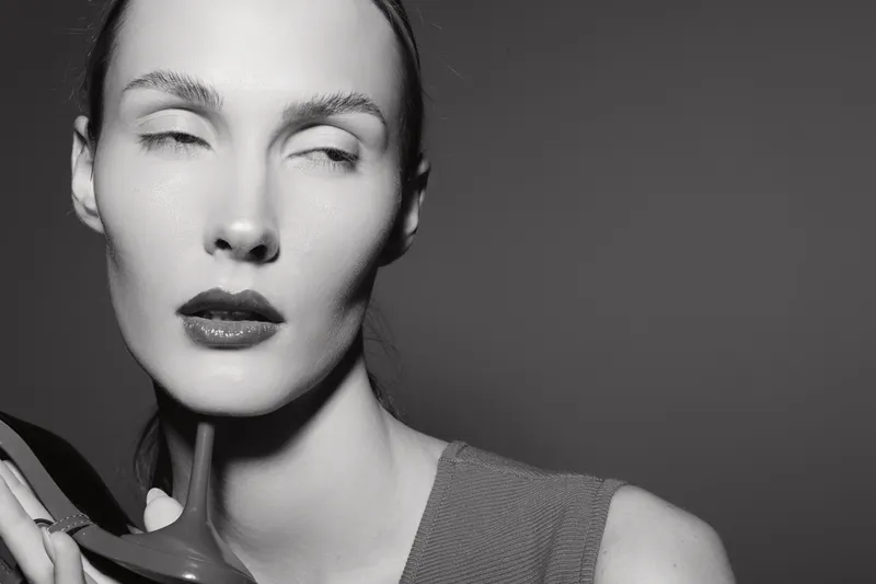 Closeup of a pale white woman with slicked back hair holding a high heel shoe near her chin, eyes closed in a studio with monochrome background, fashion concept.