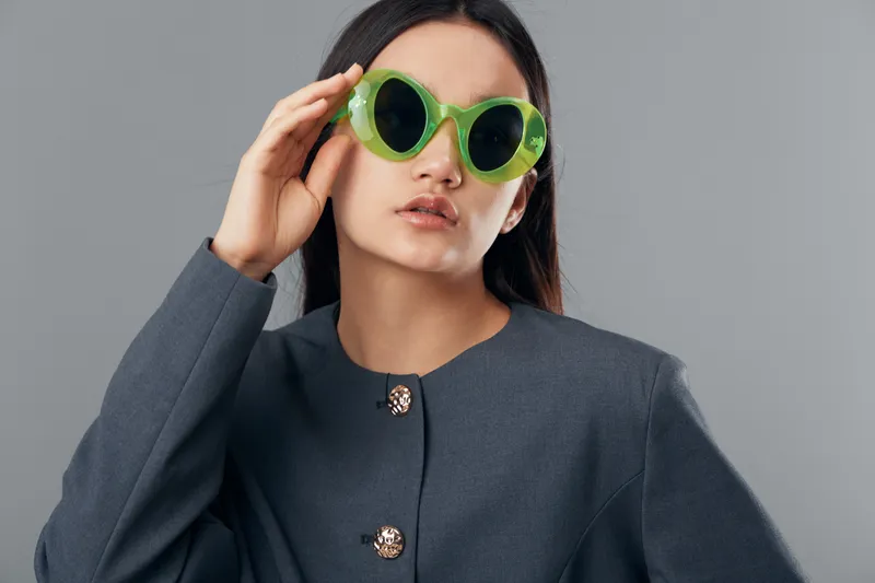 Woman with long dark hair wearing bright green sunglasses and grey buttoned coat posing against grey background in studio. Fashion, style and confident female model portrait with casual look.