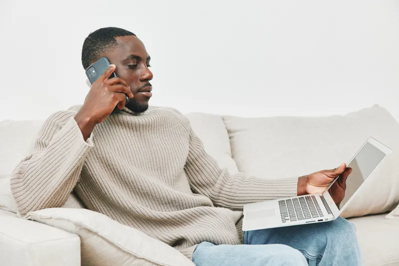 Young African man engaged in a work from home setup, speaking on the phone while using a laptop on a cozy sofa, showcasing a modern lifestyle