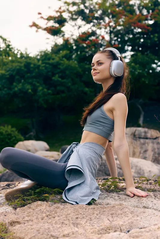 Young woman enjoying music outdoors in athletic wear, with headphones, sitting on a rock surrounded by greenery, conveying relaxation and a healthy lifestyle