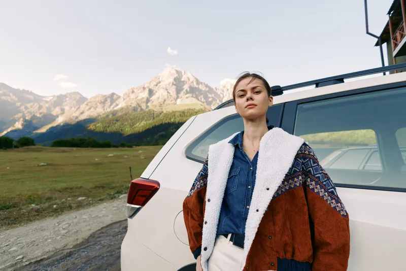 Woman standing by car with mountains behind, wearing patterned jacket and denim shirt, confident portrait during road trip travel in open landscape near SUV.