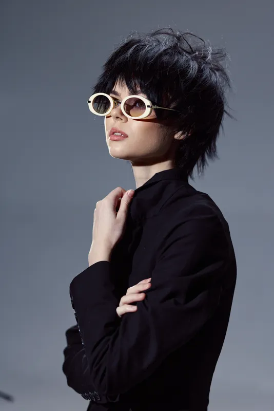 Stylish woman with short hair wearing white sunglasses and black outfit, expressing confidence in studio lighting.