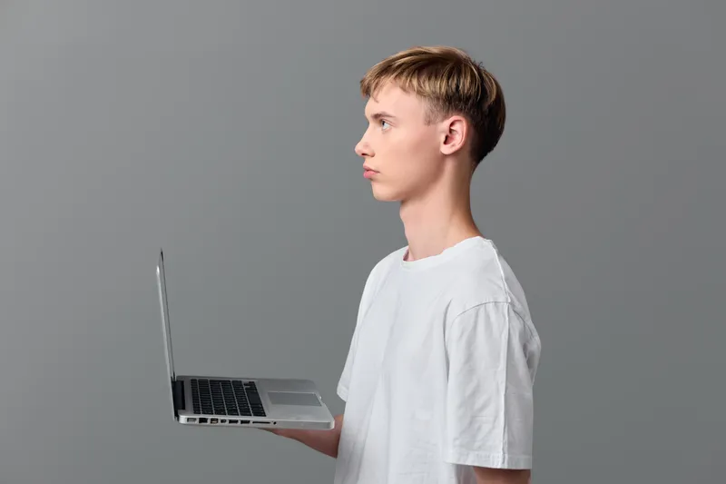 Casual young man with a laptop in profile against a gray studio backdrop, showcasing technology, education, learning, device use, screen focus, and modern digital work